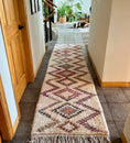 Load image into Gallery viewer, A long runner rug with a geometric diamond pattern in beige, burgundy, and brown, laid out on a tiled hallway floor. A dog is visible in the background of the hallway.
