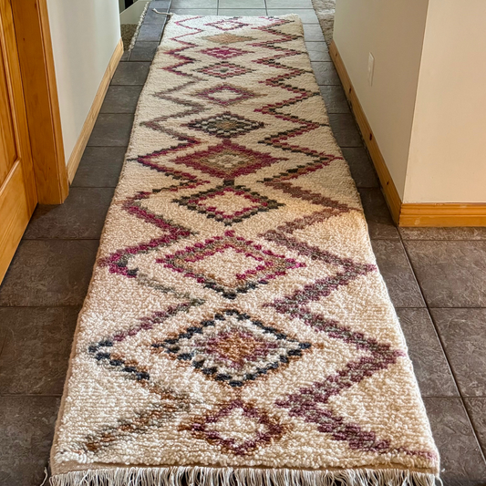 A long woven runner rug with a geometric diamond pattern in beige, brown, and muted pink, laid out on a tiled hallway floor next to a wooden door, with a dog visible in the background.